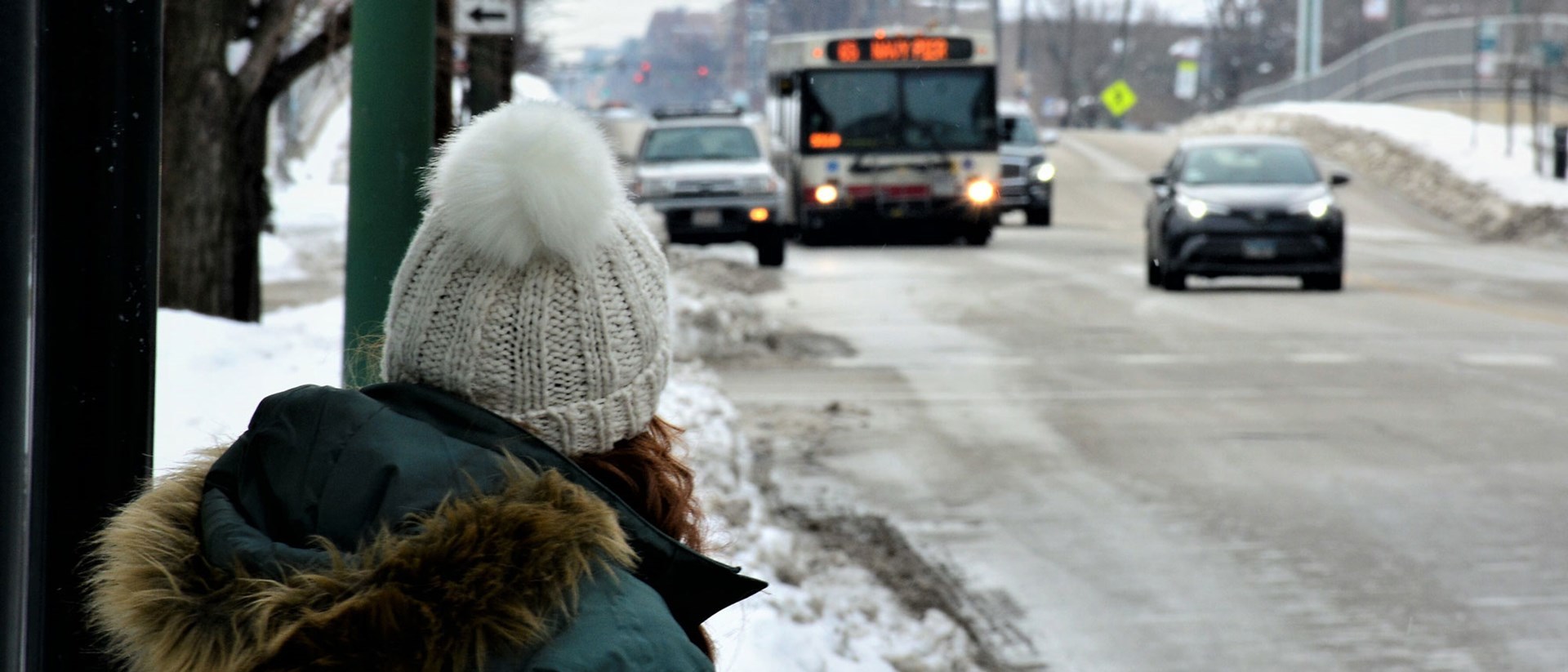 Someone in a winter hat and coat waiting at a bus stop with the bus in sight