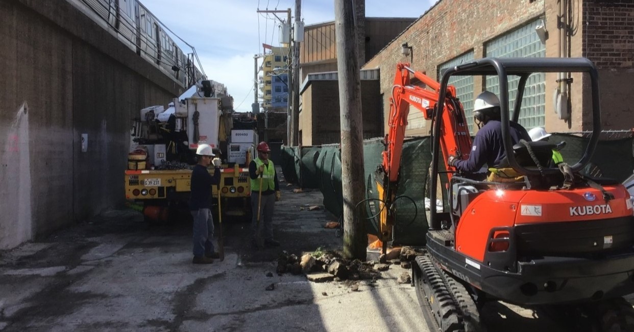 Construction crews depicted performing utilities relocation work with heavy machinery and a backhoe.